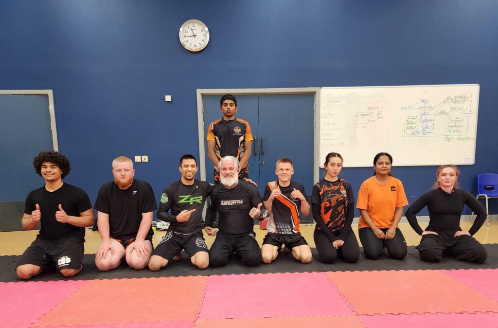 Adult NCMA grappling students kneeling on the mats after training inside the Oakgrove School sports hall.